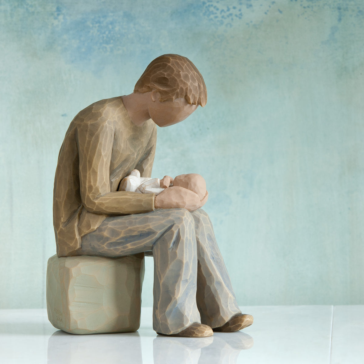 Willow Tree New Dad, 6”h hand-painted seated resin figure in cream shirt and jeans, gazing at newborn infant on lap, in front of teal painting
