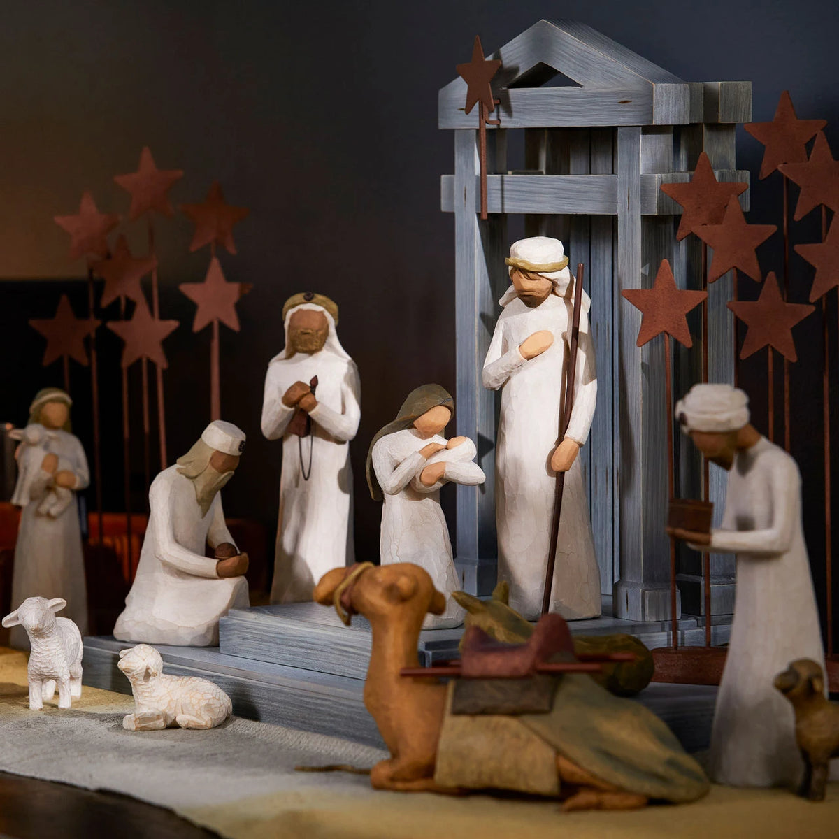 A carved nativity scene set out on a wood table laid with a linen cloth and lit candles in the background.