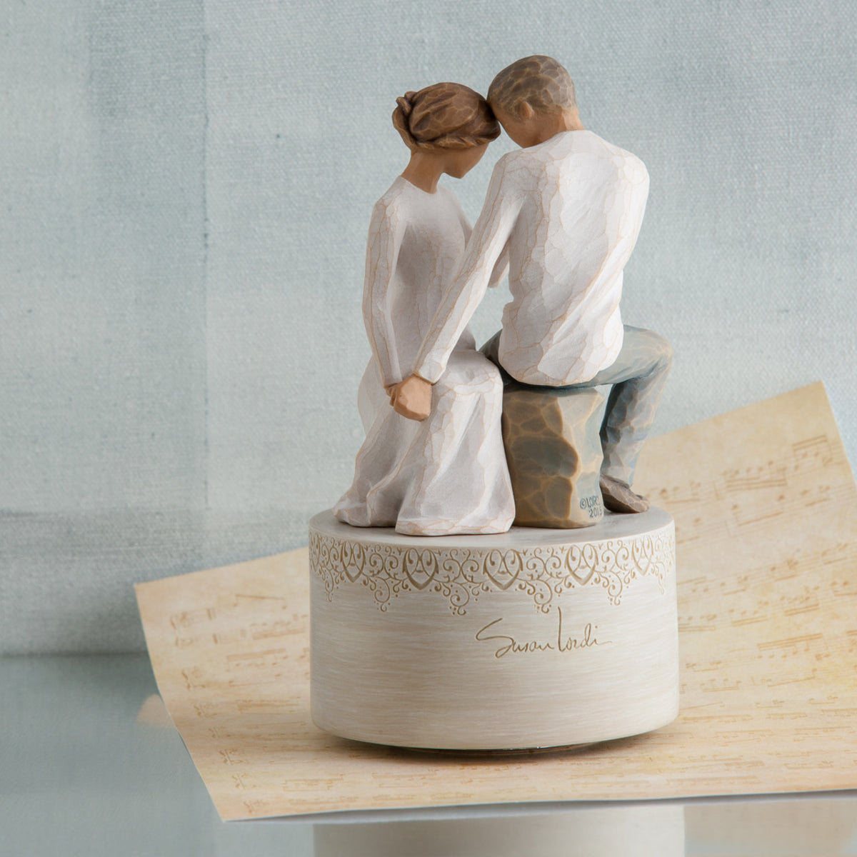 Man and woman figurine in white sitting holding hands on round large plaque and tan paper below it