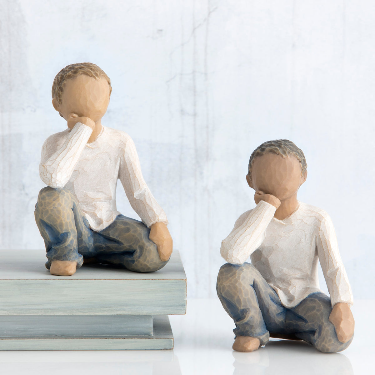 Two young brunette boy figurines in white and jeans sitting down with one arm to their face next to stack of books