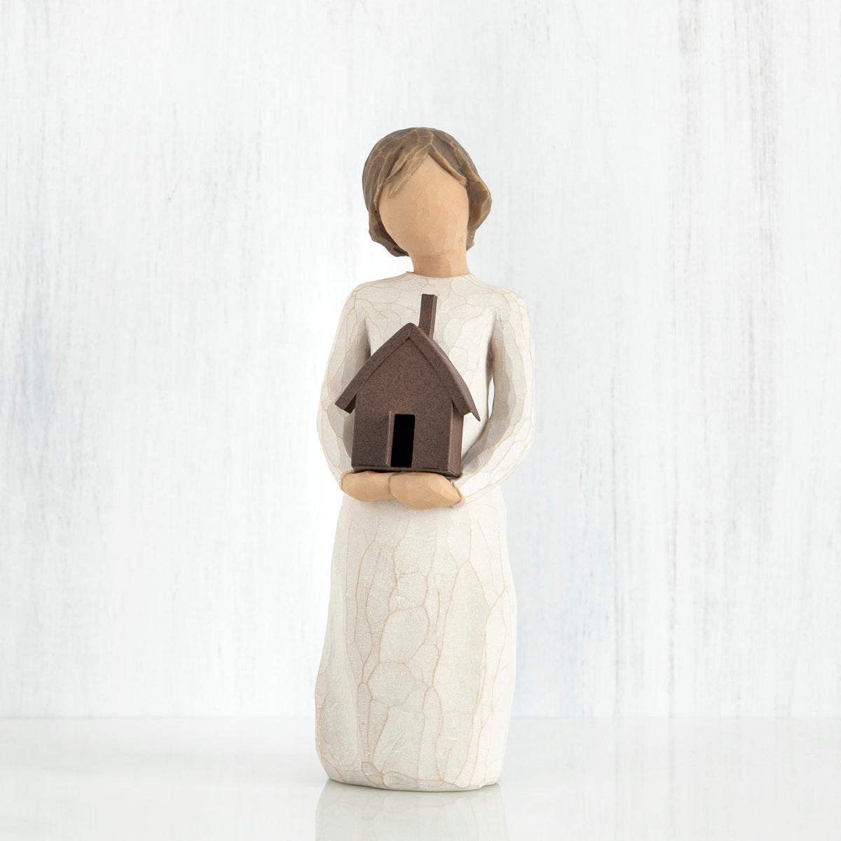 Faceless girl figurine facing forward wearing white dress, holding brown house figurine in her arms