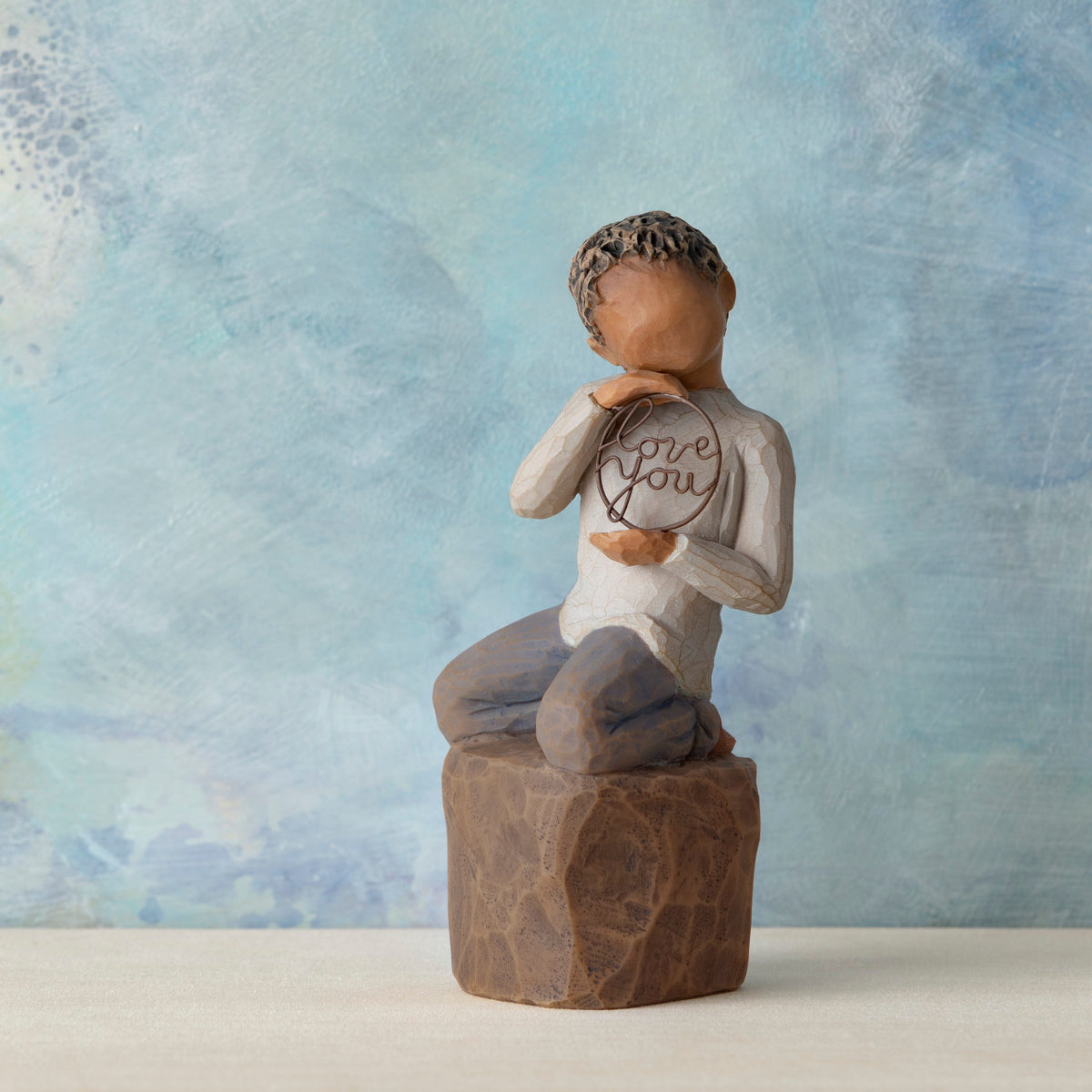 Willow Tree Love you too (darker skin), 4.5”h hand-painted resin figure of young child in cream shirt and jeans, kneeling on rock, holding wire circle with ‘love you’ inside, in front of blue painting