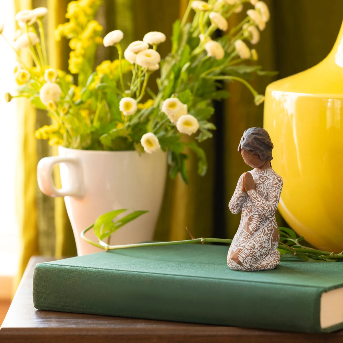 Willow Tree Prayer of Peace, 4”h hand-painted kneeling figure with darker skin and hair, in cream dress carved with nature imagery, in prayer/meditation pose, on desk with yellow florals