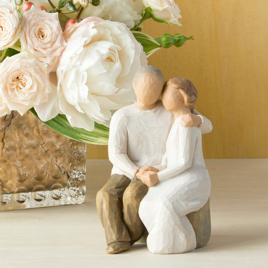 Willow Tree Anniversary, 6”h hand-painted resin figure of older couple sitting on bench holding hands, both in neutral clothing, on table with white roses