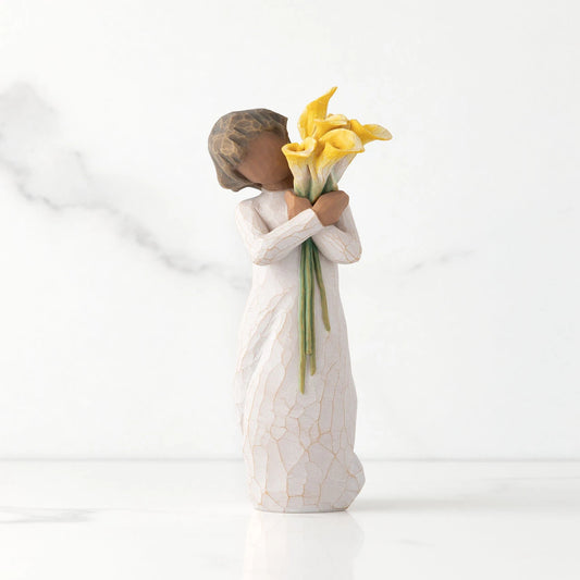 Front view of standing figure in cream dress with darker skin holding yellow calla lilies in her arms