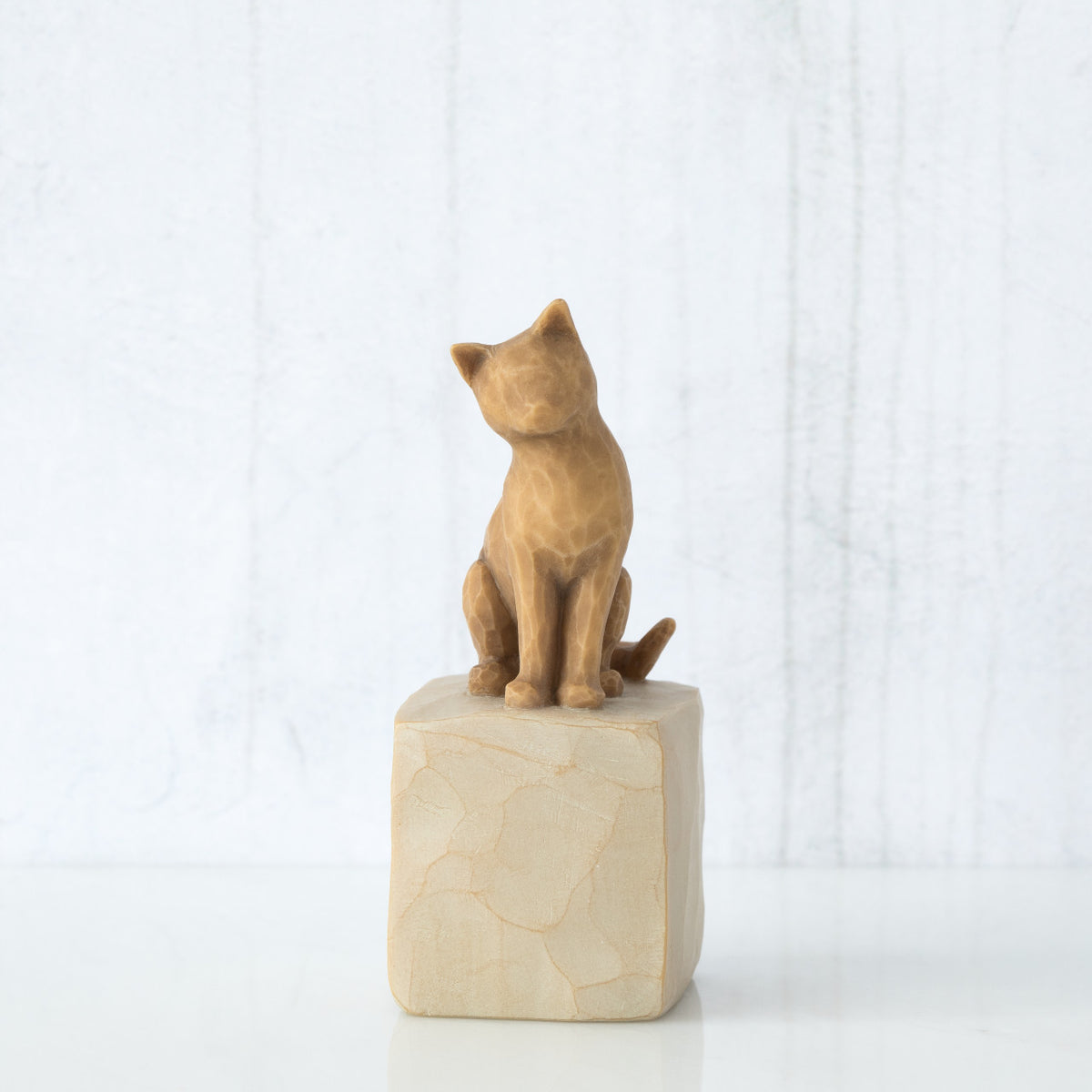 Figure of orange cat with tilted head perched atop a cream colored rectangular pedestal, facing front