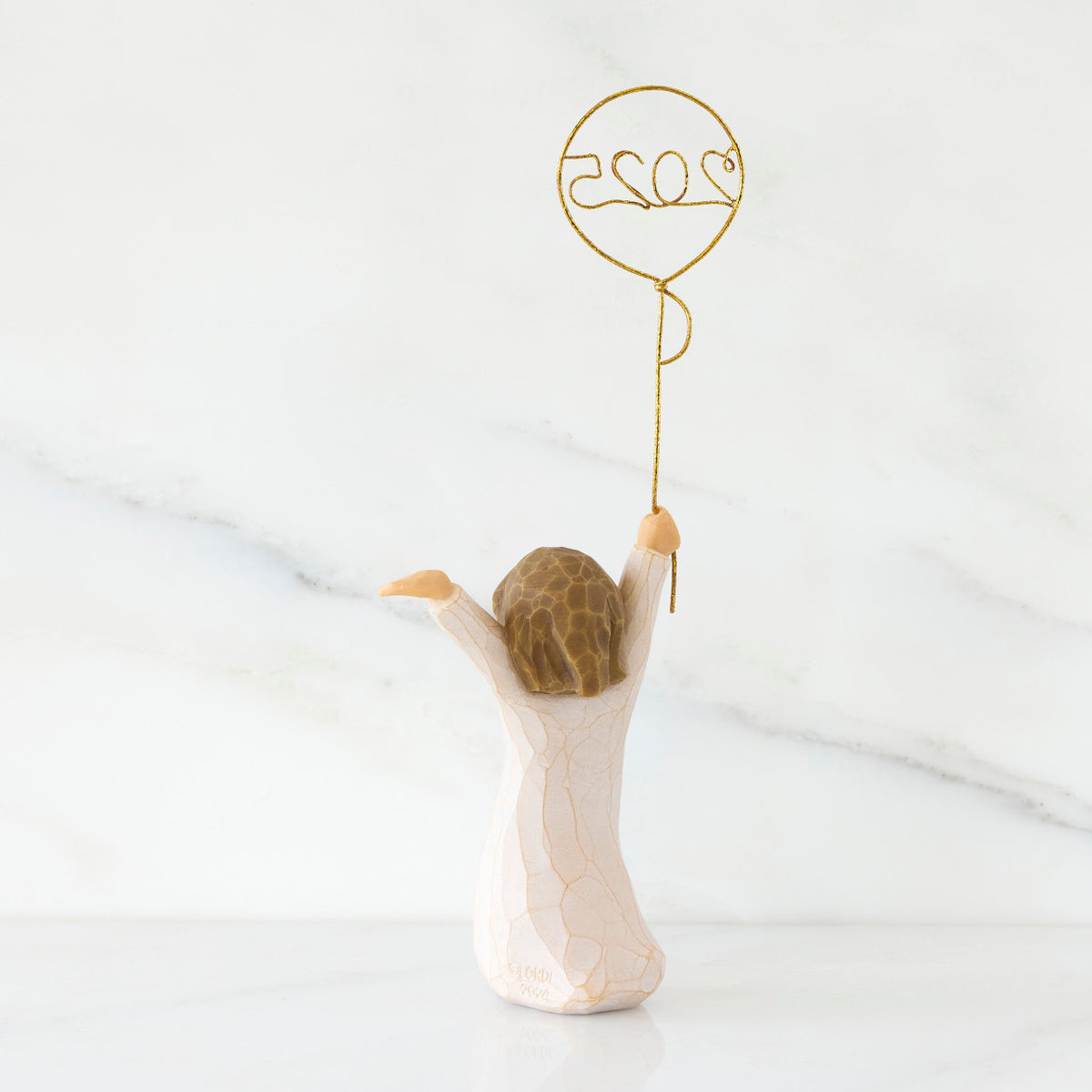 Back view: standing figure in cream dress, arms raised, holding gold wire balloon with 2025 written in gold wire inside