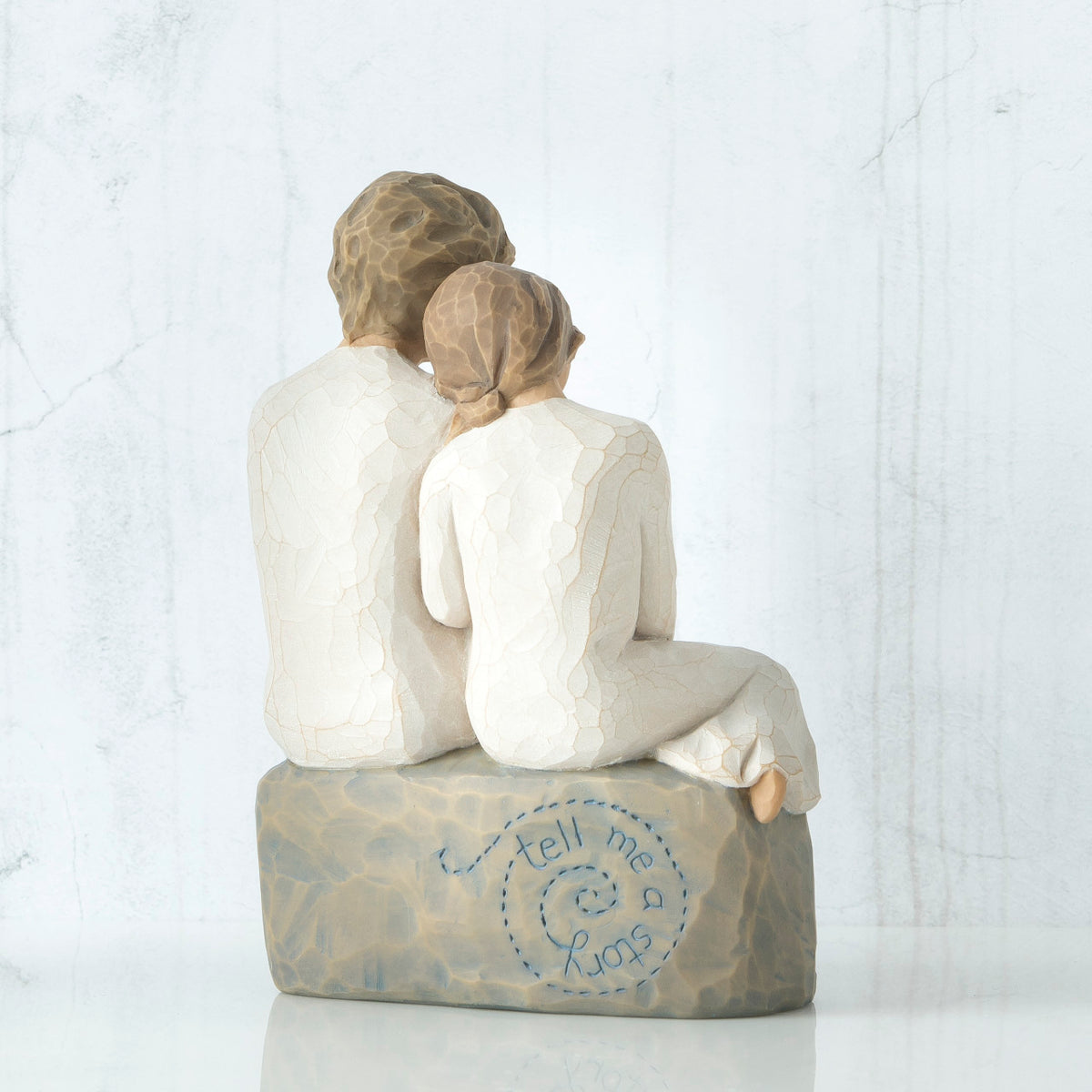 Back view of boy and girl figurine in white sitting on grey rock that says 'tell me a story' in blue carved in